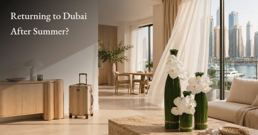 Living room in a Dubai apartment overlooking the marina, styled with white orchid arrangements in tall vases beside a packed suitcase, welcoming the return to fresh flowers after summer