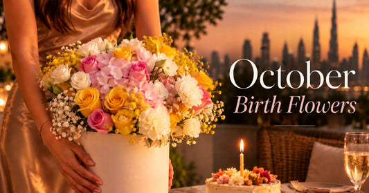 Woman holding a pink, yellow and white October birthday flower box beside a candlelit birthday cake on a Dubai terrace at sunset, AIWA Flowers