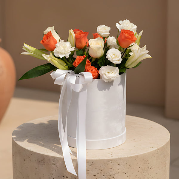 White velvet round box arrangement with orange and white roses, white lilies, carnations, and lisianthus