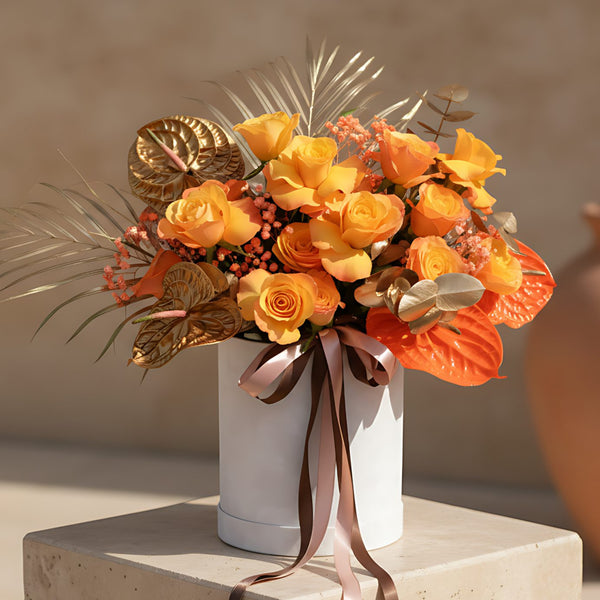 White velvet box arrangement with orange roses, gold and orange anthuriums, gold foliage, and orange gypsophila