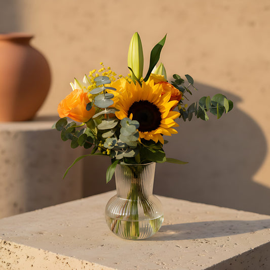 Clear glass vase arrangement with yellow and orange roses, sunflower, white lily, gypsophila, and eucalyptus