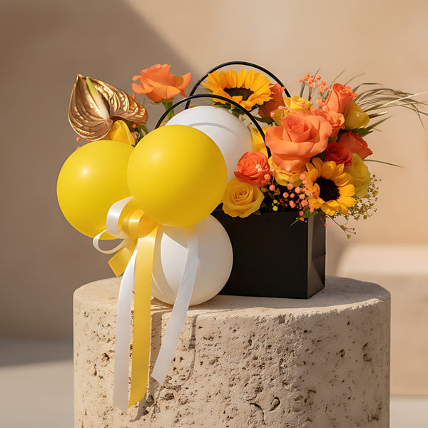 Bag-box floral arrangement with yellow and white balloons, orange and yellow roses, sunflowers, and gold accents