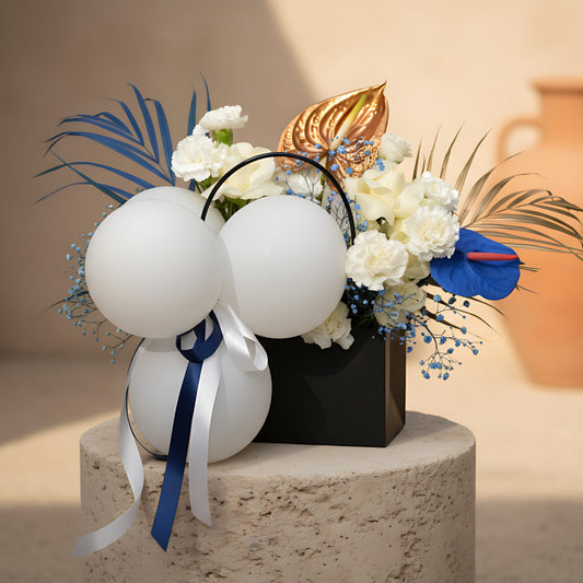 Bag-box floral arrangement with white roses, white carnations, blue gypsophila, balloons, and blue and gold accents