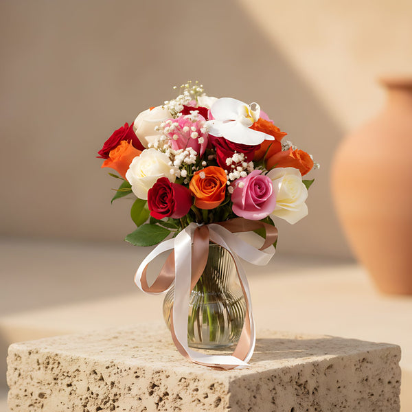 Clear glass vase arrangement with red, pink, and orange roses, gypsophila, and a white orchid