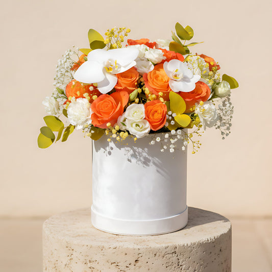 White velvet box arrangement with orange roses, white carnations, lisianthus, orchids, eucalyptus, and gypsophila