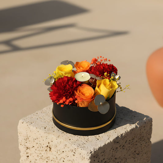 Black box floral arrangement with yellow and orange roses, carnations, gypsophila, gold eucalyptus, and a candle