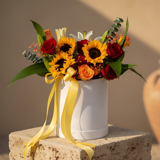 White velvet round box arrangement with sunflowers, orange and red roses, lilies, gypsophila, and eucalyptus