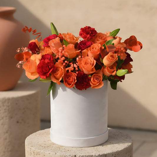 White velvet box arrangement with orange and red roses, red carnations, white lilies, and eucalyptus