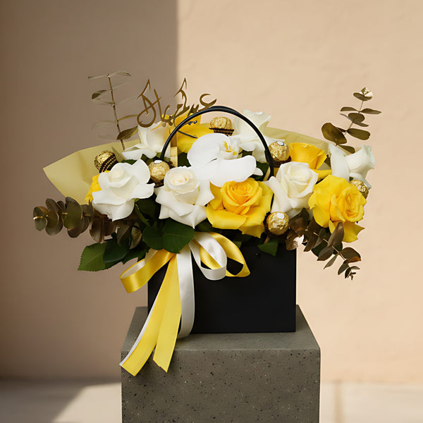 Al Adha Blooms floral arrangement with yellow and white roses, orchid, Ferrero chocolates, and Arabic topper in black bag-box by Aiwa Flowers Dubai