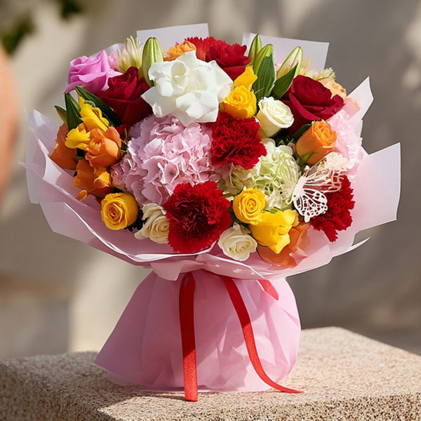 Hand-tied bouquet of mixed color roses with hydrangeas, lilies, red carnations, and a butterfly accent in soft pink wrapping by Aiwa Flowers Dubai

