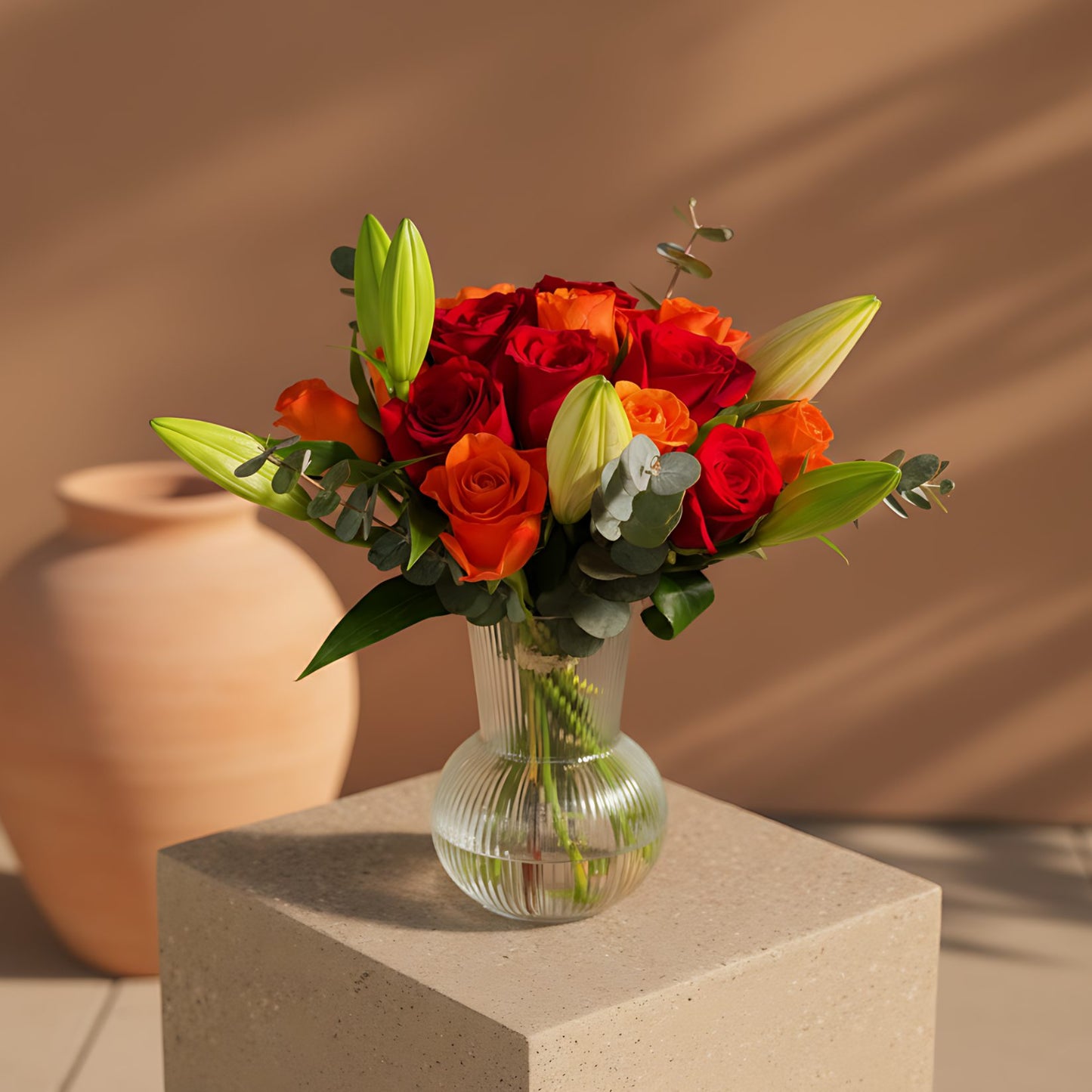 Stella floral arrangement with orange and red roses, lilies, and eucalyptus in a clear glass vase by Aiwa Flowers Dubai