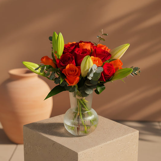 Stella floral arrangement with orange and red roses, lilies, and eucalyptus in a clear glass vase by Aiwa Flowers Dubai
