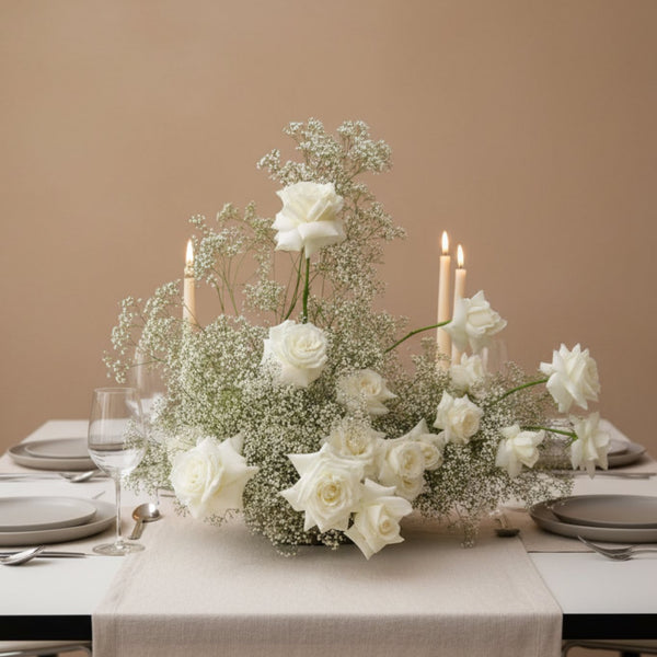 Elegant ivory rose and gypsophila floral centerpiece with candles arranged on a refined dining table.