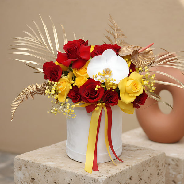White velvet box arrangement with red and yellow roses, white orchid, yellow gypsophila, and gold accents