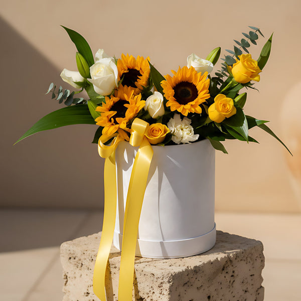 White velvet round box arrangement with sunflowers, yellow and white roses, lilies, carnations, and eucalyptus