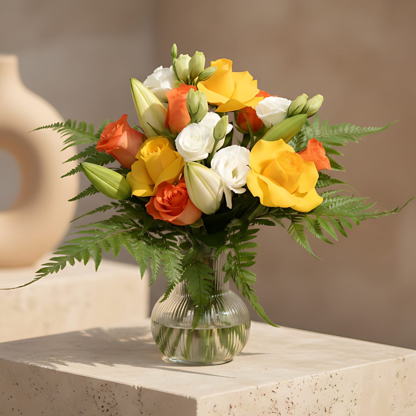 Clear glass vase arrangement with orange and yellow roses, white lilies, lisianthus, and fern leaves