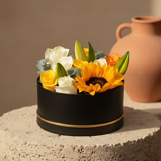 Black round box flower arrangement with sunflower, mixed white, orange and yellow roses, lilies, and eucalyptus
