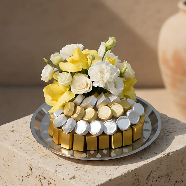 Elegant silver plate arrangement with yellow and white roses, white carnations, lisianthus, and gourmet chocolates