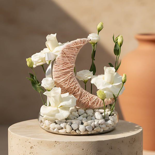Elegant Ramadan floral arrangement with white roses and white lisianthus, accented by a pink knotted moon detail and white pebbles on a glass tray, available for delivery in Dubai.
