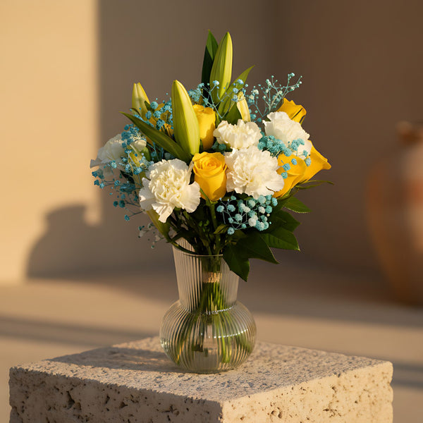 Laura floral arrangement with yellow roses, white carnations, white lilies and blue gypsophila in a clear vase by Aiwa Flowers