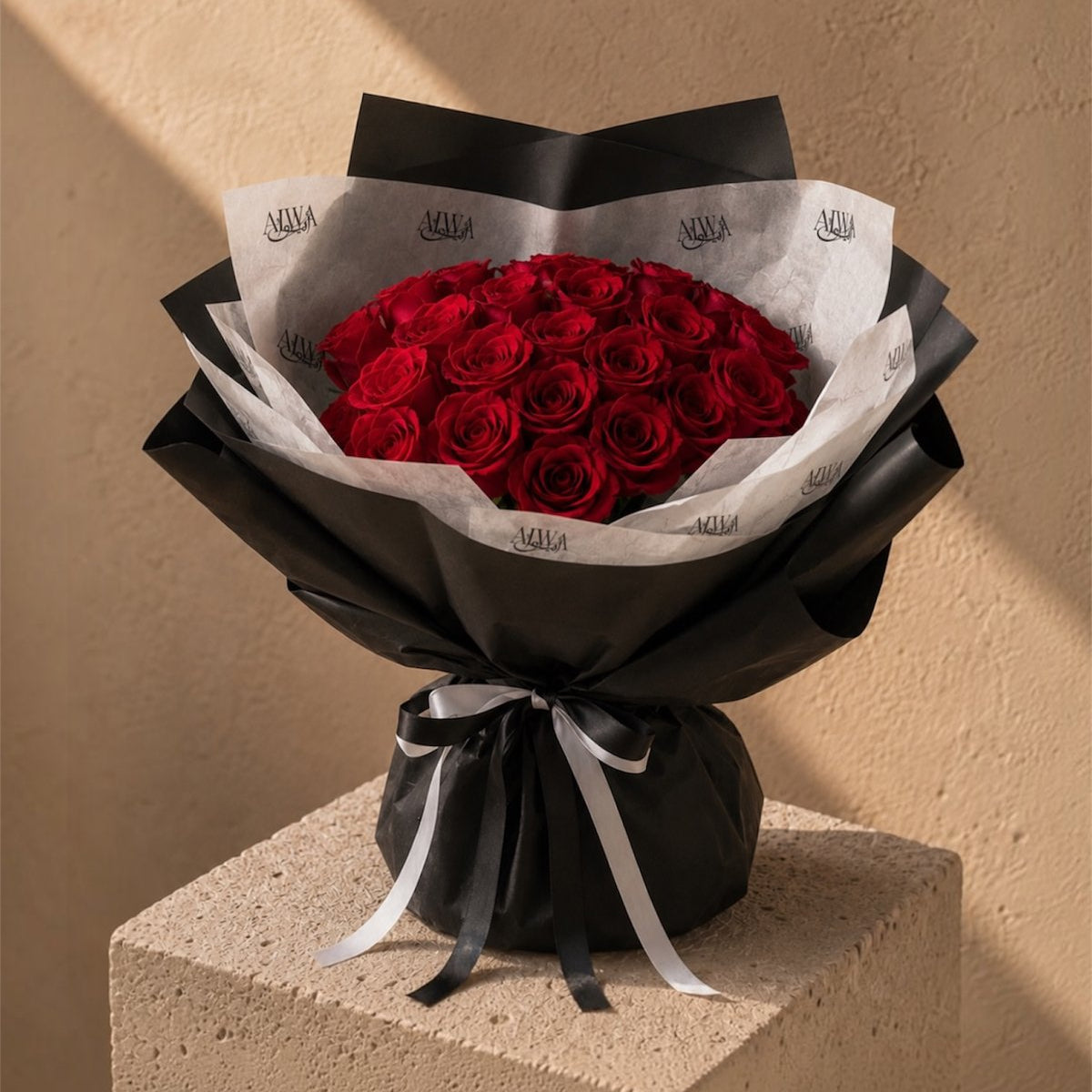 A bouquet of red roses wrapped in black paper and white tissue paper with a black and white ribbon, displayed on a textured beige pedestal against a beige wall.