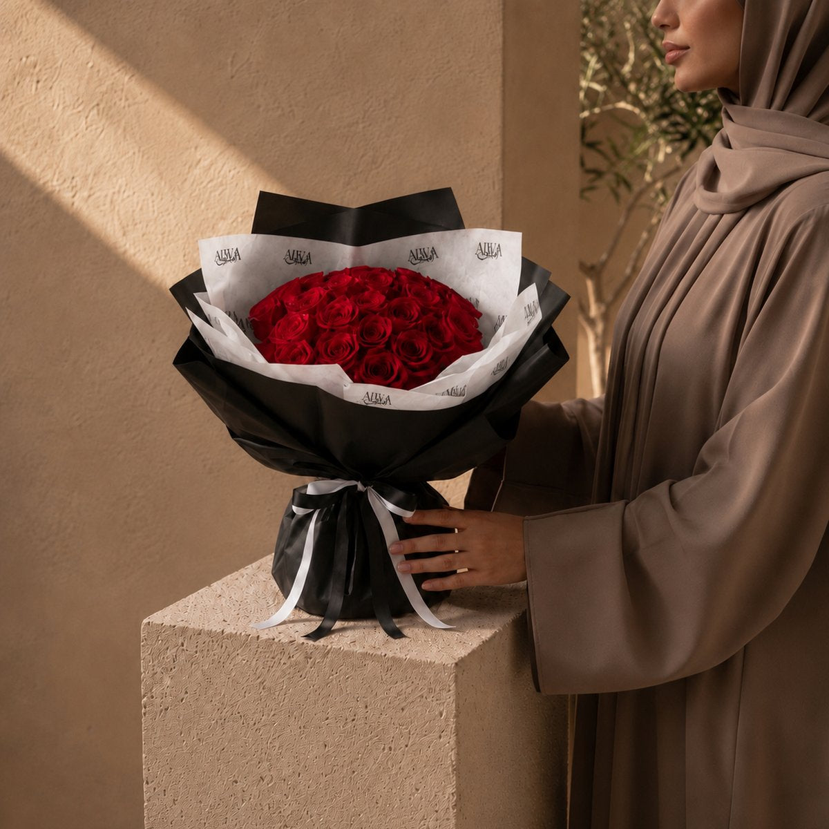 A bouquet of red roses wrapped in black paper and white tissue paper with a black ribbon, displayed on a beige stone pedestal next to a woman wearing a brown abaya and hijab.