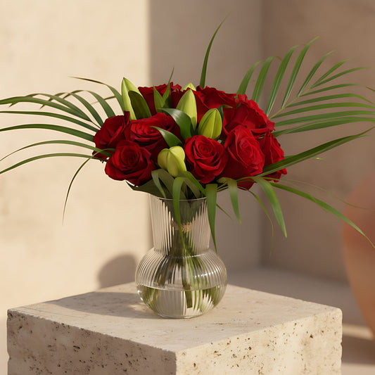 Nora arrangement with red roses, lilies and areca leaves in a clear glass vase
